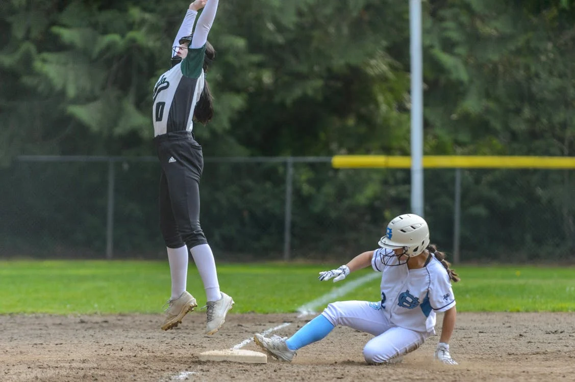 Two female softball players in action as a runner slides into second base while the shortstop leaps for the throw.